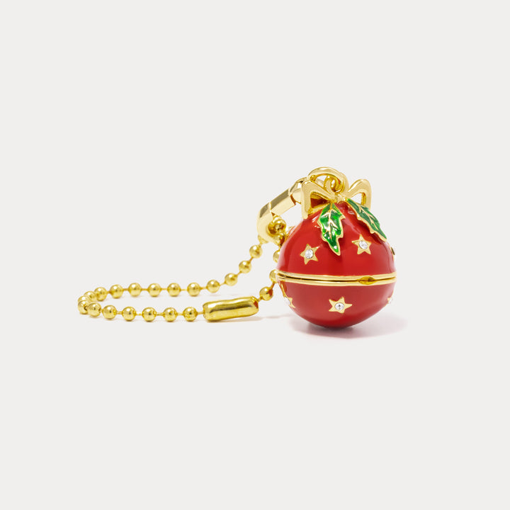 A red Christmas ornament keychain with gold-tone metal details, featuring a bow, holly leaves, and stars, attached to a gold ball chain on a light gray background.